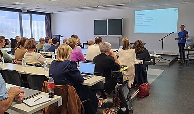 people sitting at desks listening and taking notes of the presentation with presenter standing in frontm und eine Person steht vorne und präsentiert